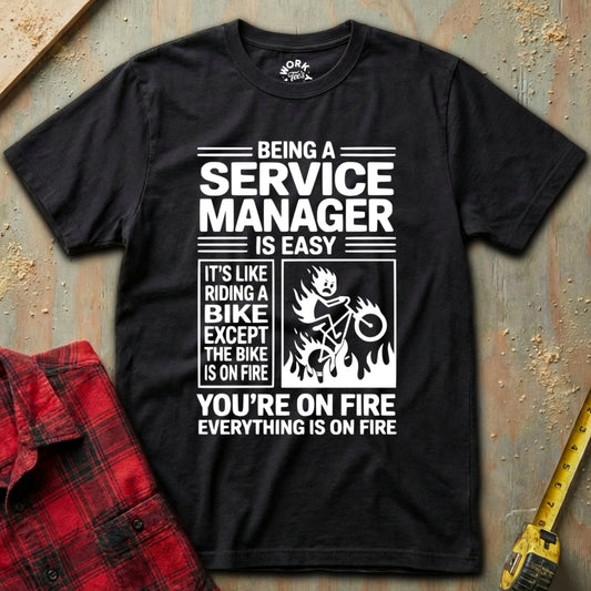 Black t-shirt with humorous text design on a textured surface with a red plaid shirt and yellow tool in the background.