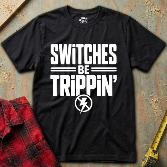 Black t-shirt with 'SWITCHES BE TRIPPIN'' text on a textured surface with a red plaid shirt and yellow tool in the background.