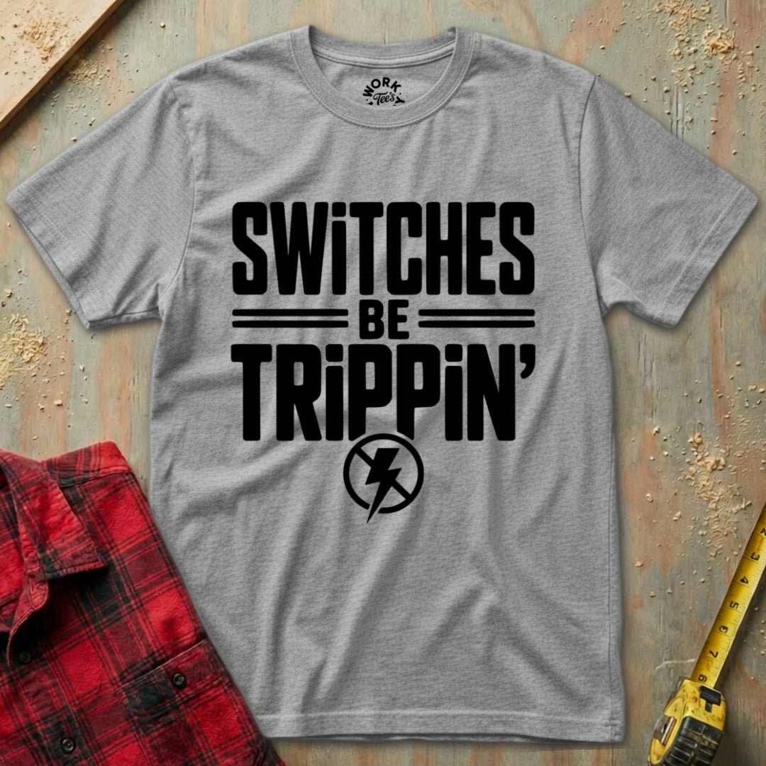 Gray t-shirt with 'SWITCHES BE TRIPPIN'' text on a textured surface with a red plaid shirt and yellow tool in the background.