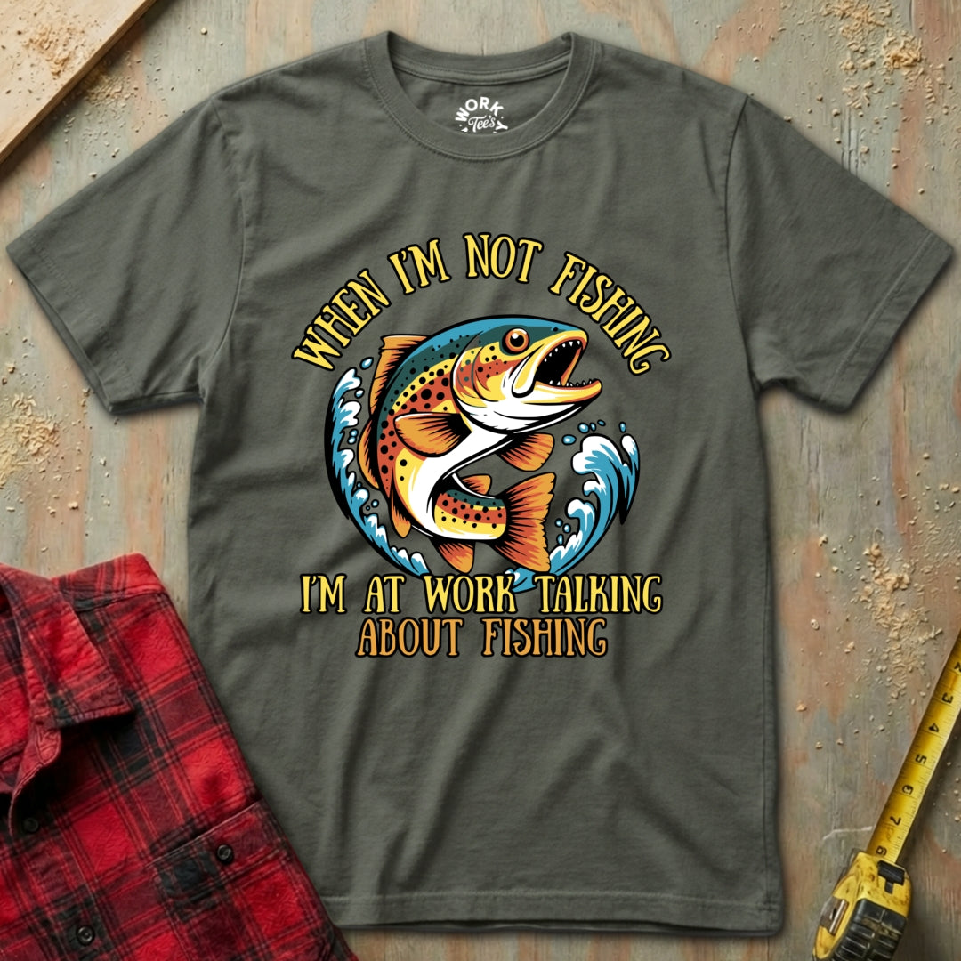 Green t-shirt with fishing graphic and text on a wooden surface with plaid shirt and tape measure.