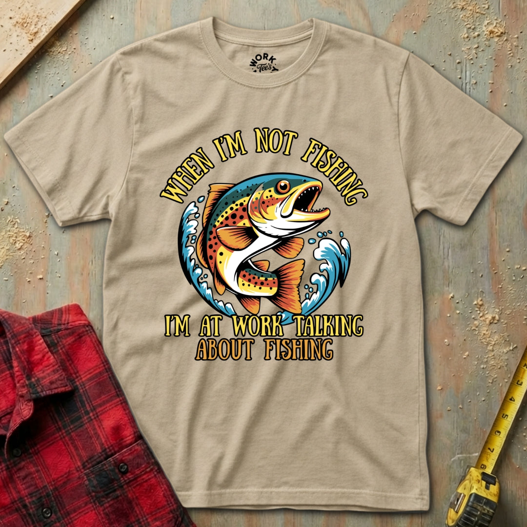 Beige t-shirt with fishing graphic and text on a wooden surface with plaid shirt and tape measure.