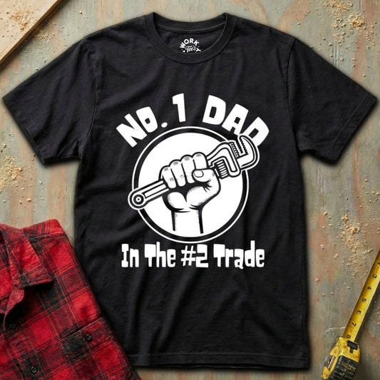 Black t-shirt with 'No. 1 Dad in the #2 Trade' graphic on a textured surface with a red plaid shirt and yellow tool in the background.