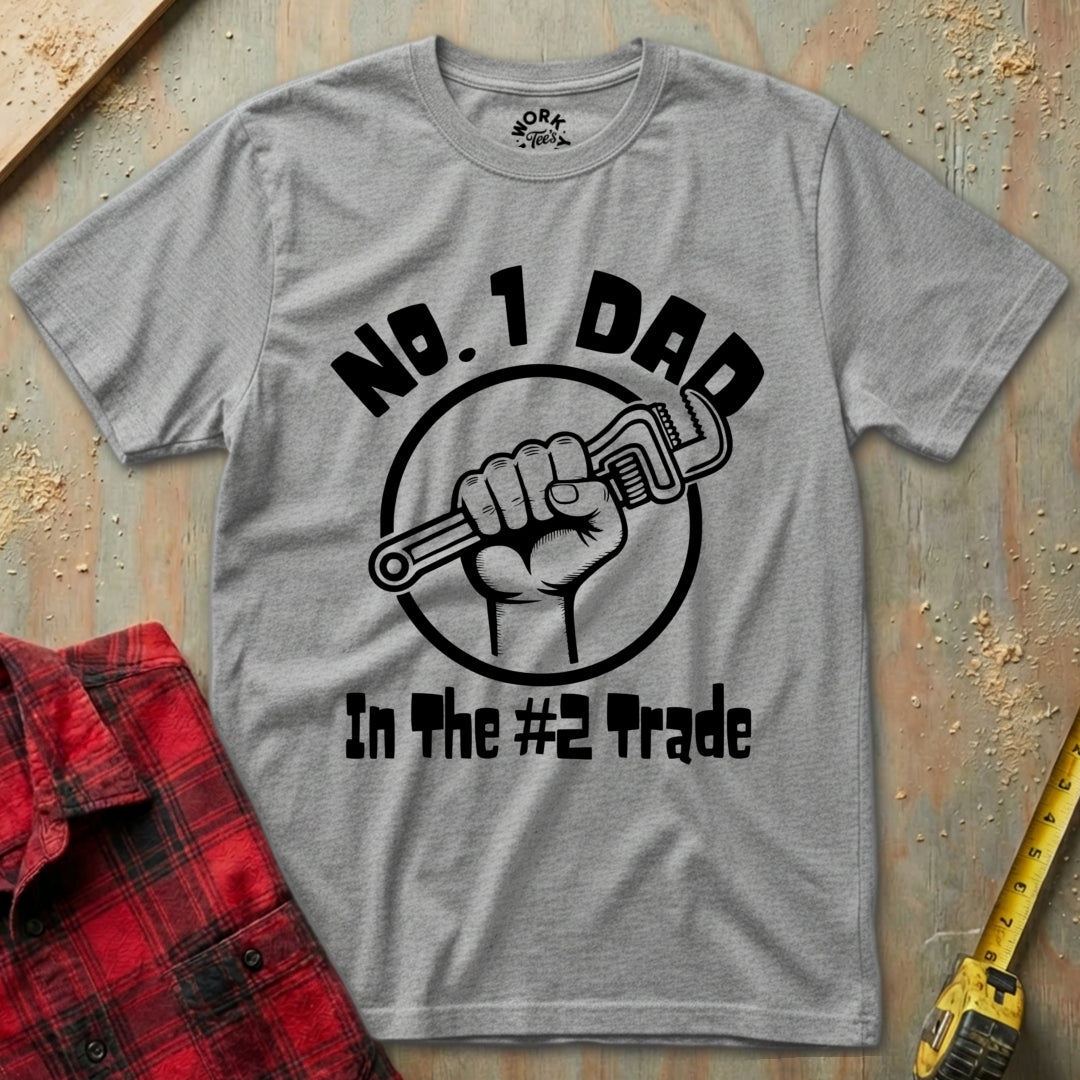 Gray t-shirt with 'No. 1 Dad in the #2 Trade' graphic on a wooden surface with a red plaid shirt and yellow tool in the background.